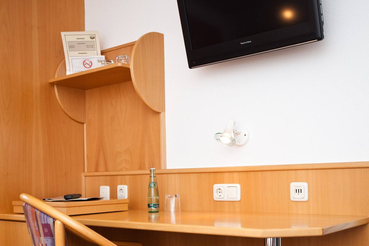 A hotel room with a wooden desk, a chair, a flat-screen TV mounted on the wall, and a shelf with a bottle and glasses.