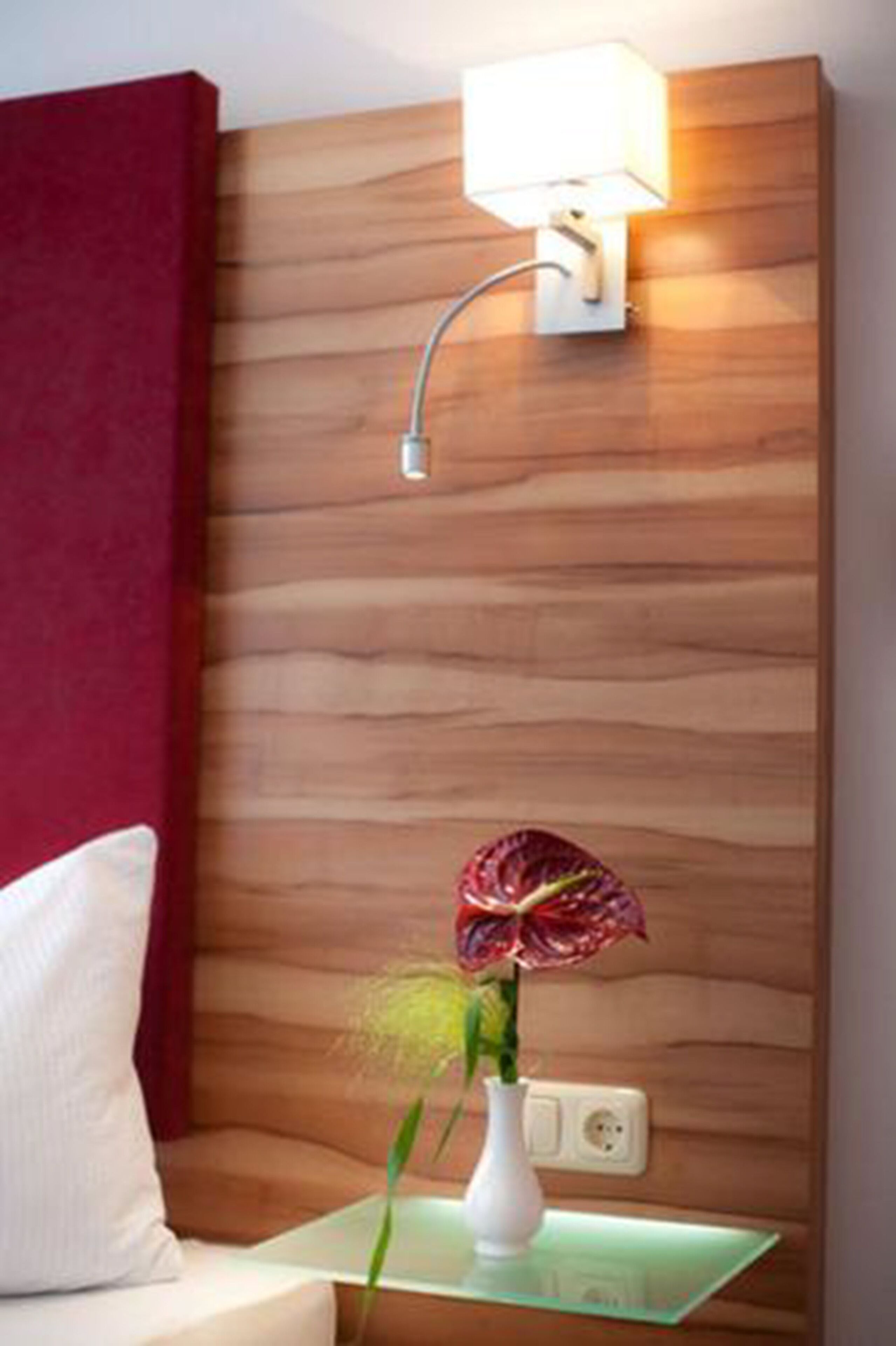 A room with a wooden headboard, a bedside table with a lamp, a vase with a plant, and a red curtain.