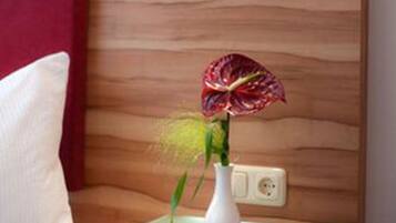 A room with a wooden headboard, a bedside table with a lamp, a vase with a plant, and a red curtain.