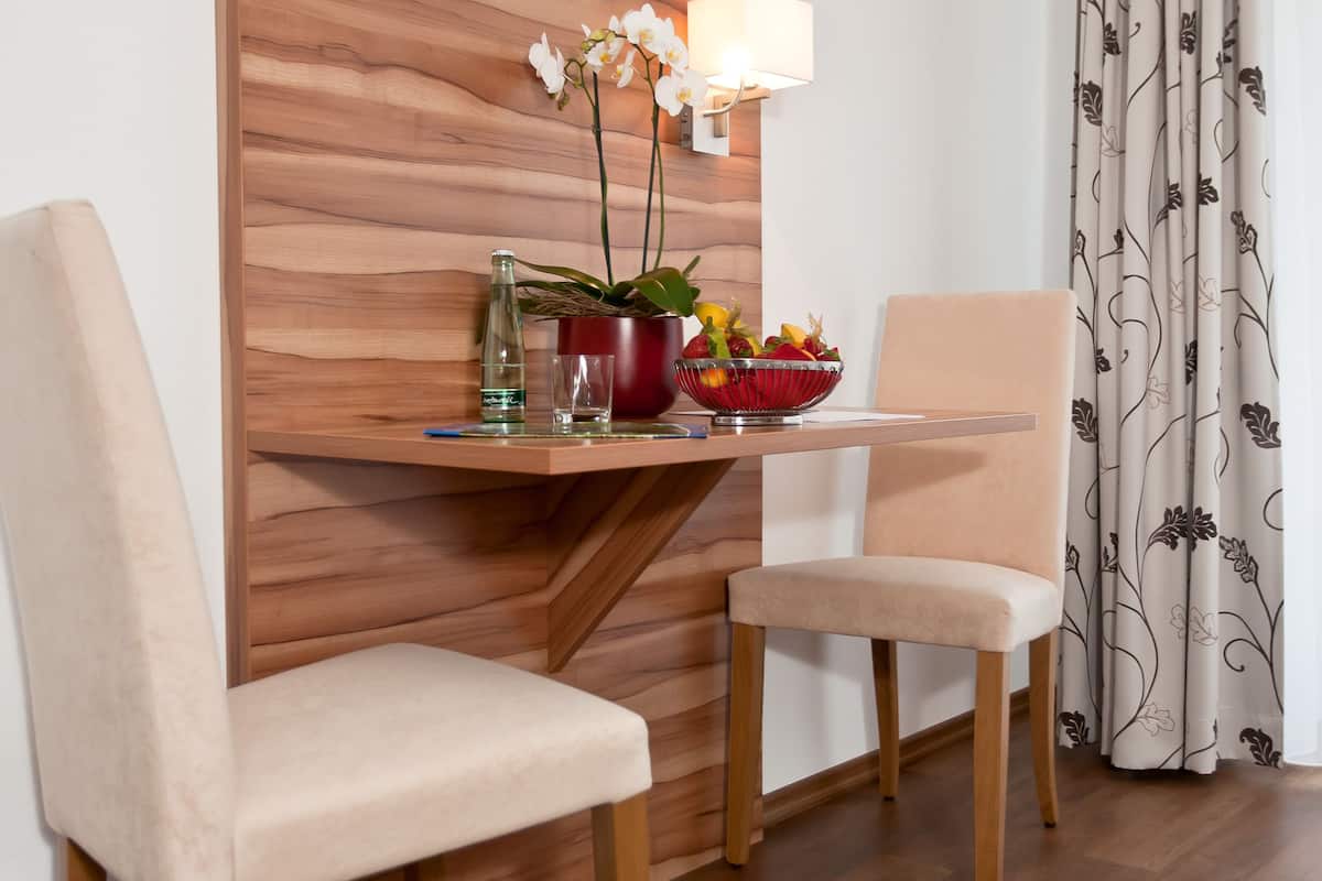 A modern room with a flat-screen TV mounted on a wooden wall, two upholstered chairs, a small table with a vase of flowers, and a patterned curtain.