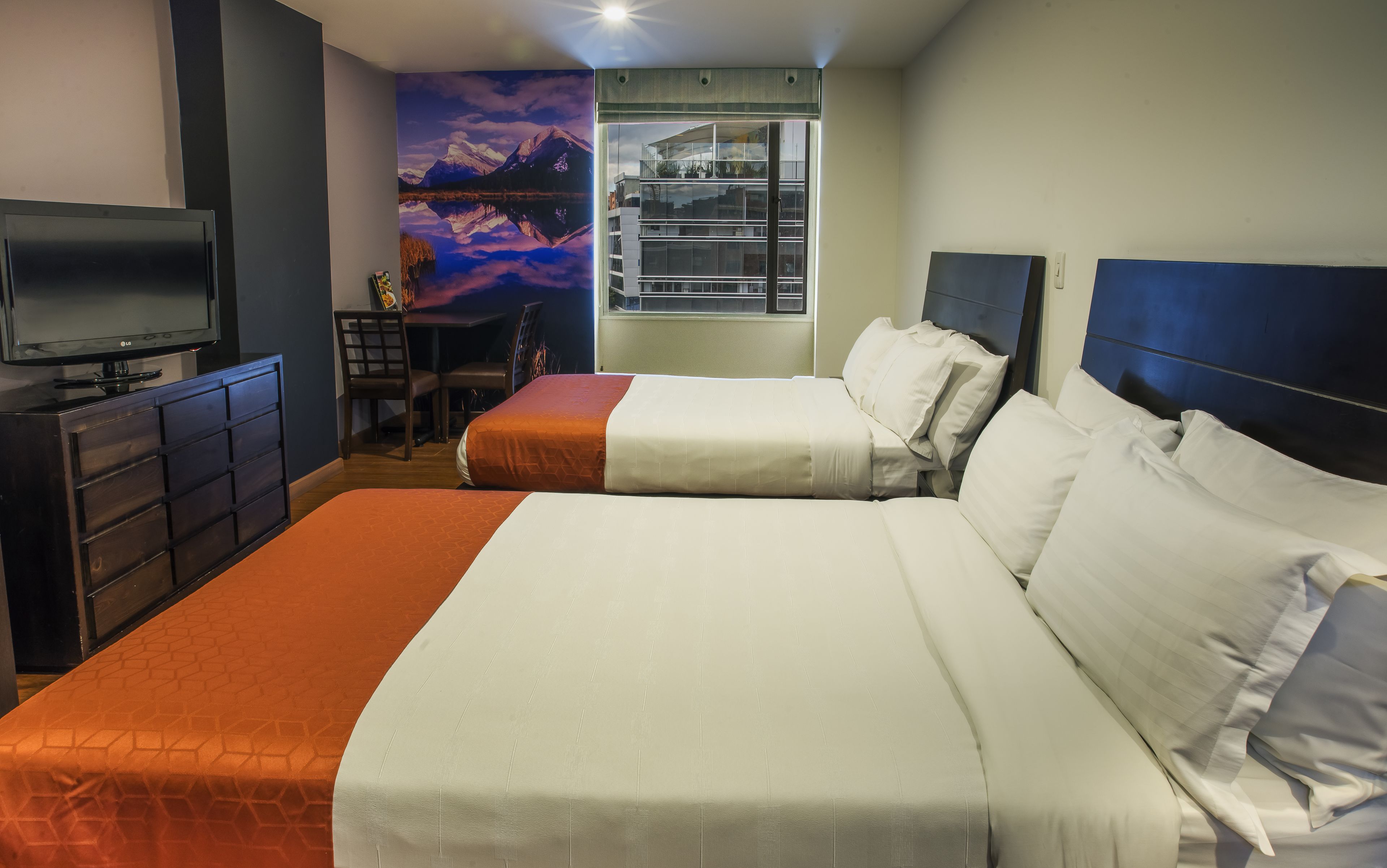 Habitación de hotel con dos camas, televisión, escritorio y un mural de montañas.