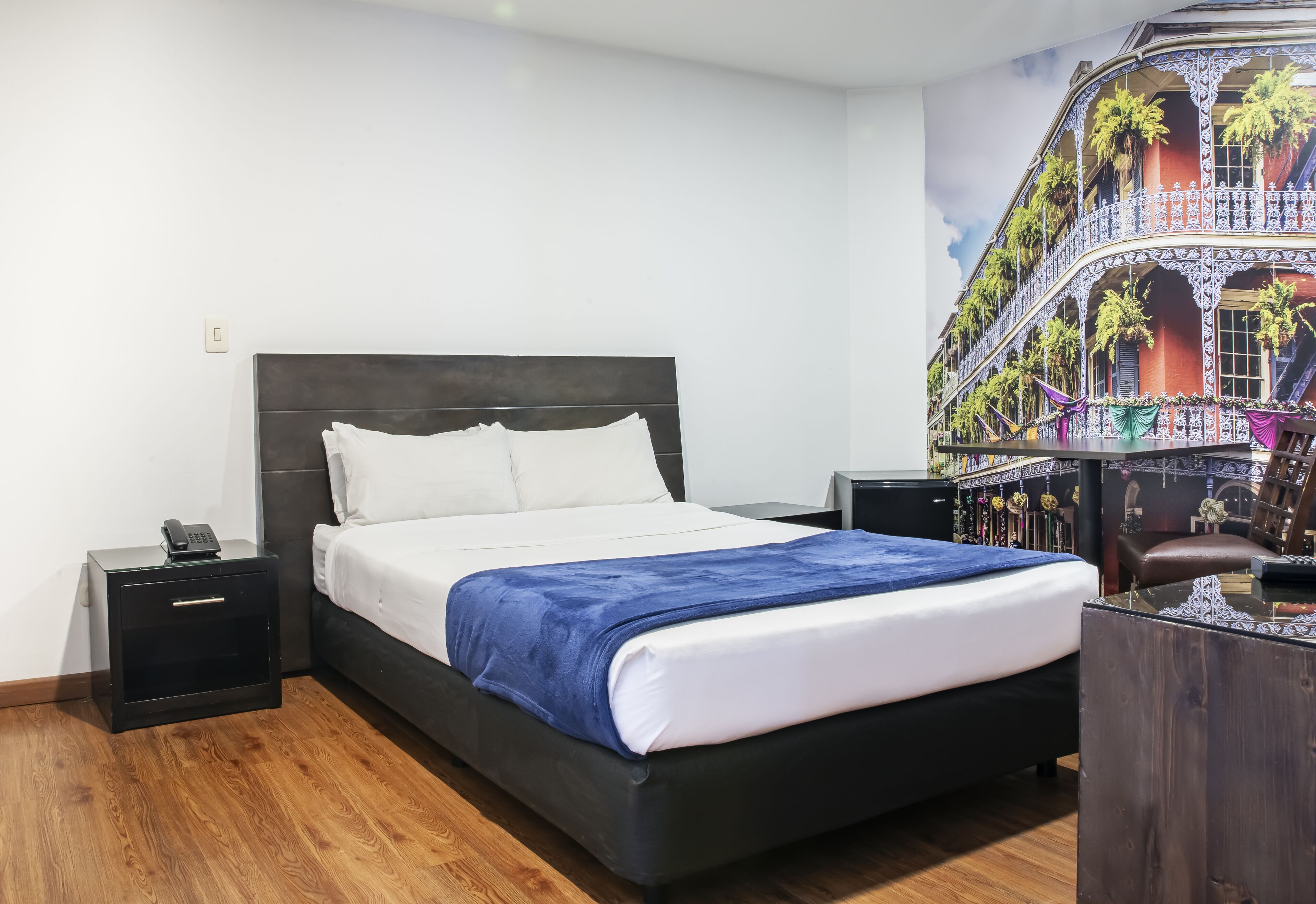 A hotel room with a bed, bedside tables, a nightstand, a desk, and a large window with a view of a building.