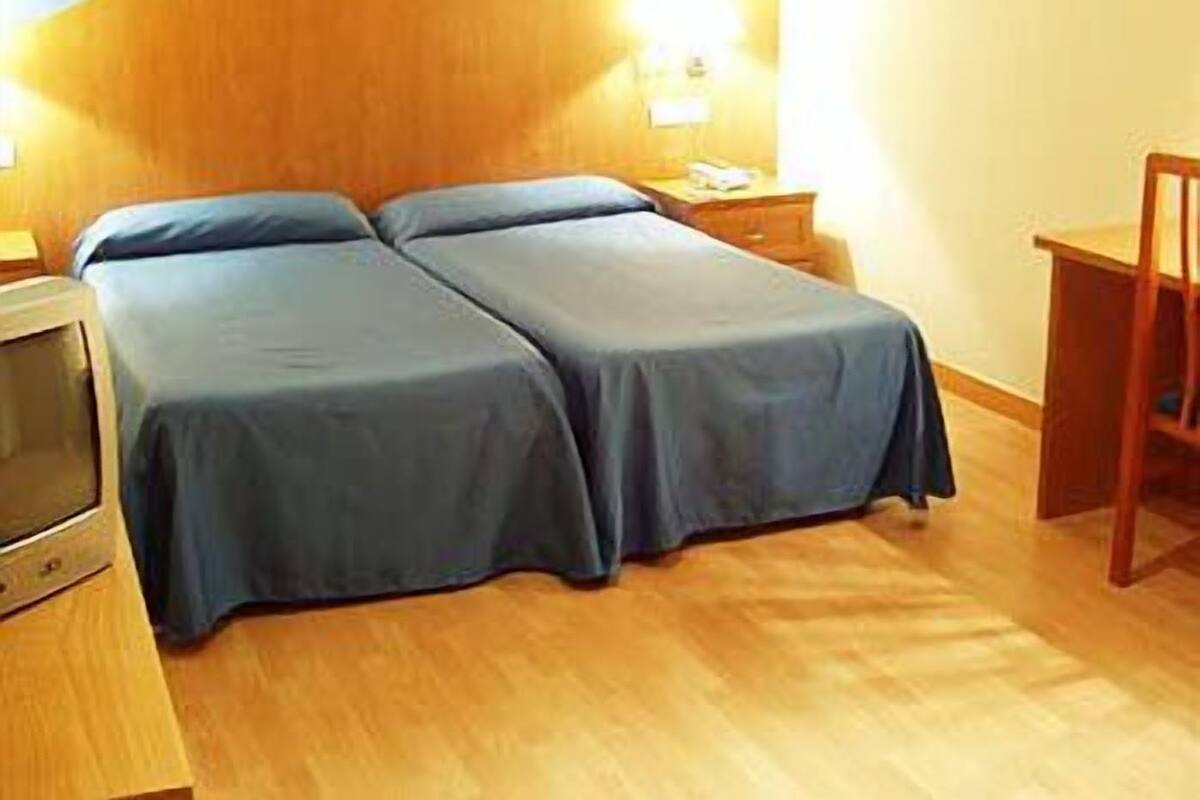 A room with two beds, a wooden floor, a desk with a chair, and a wall-mounted light.