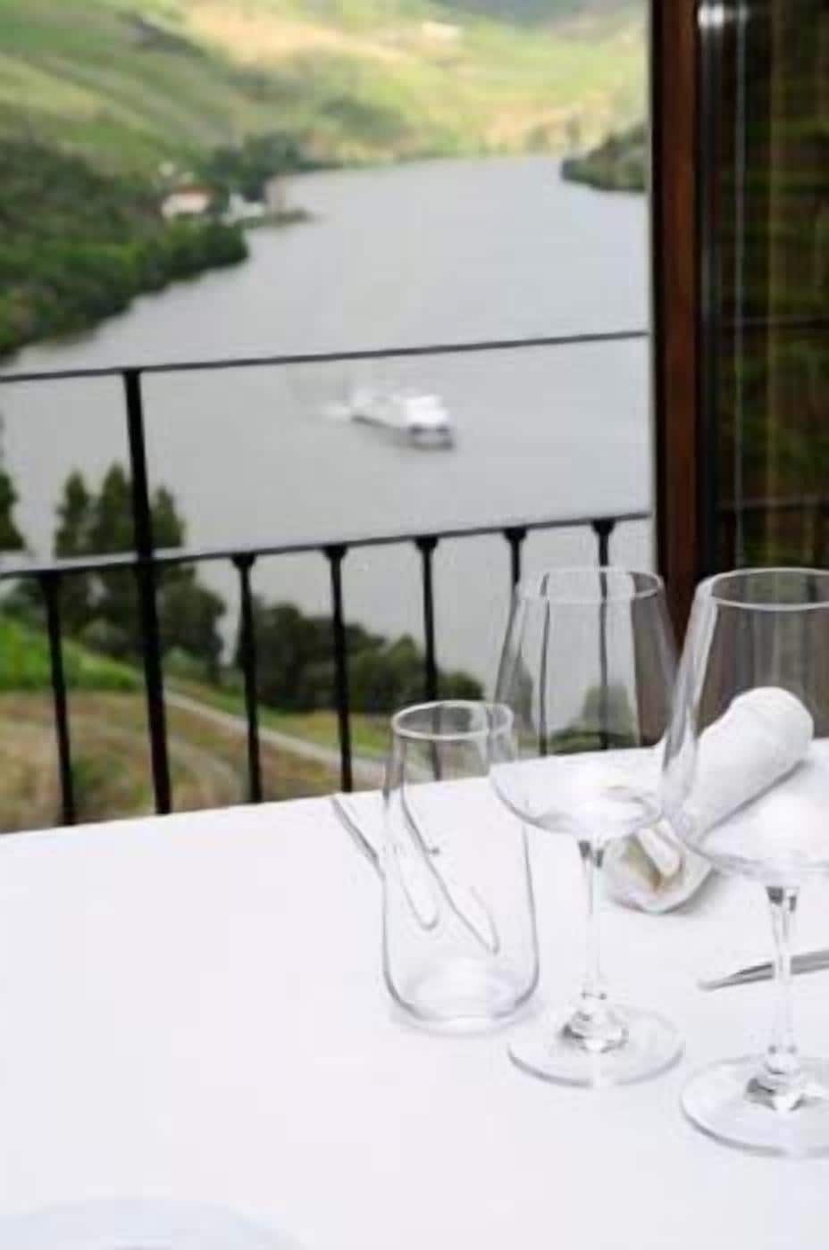 A table with three wine glasses and a water glass, set against a backdrop of a scenic view of a river and a boat.