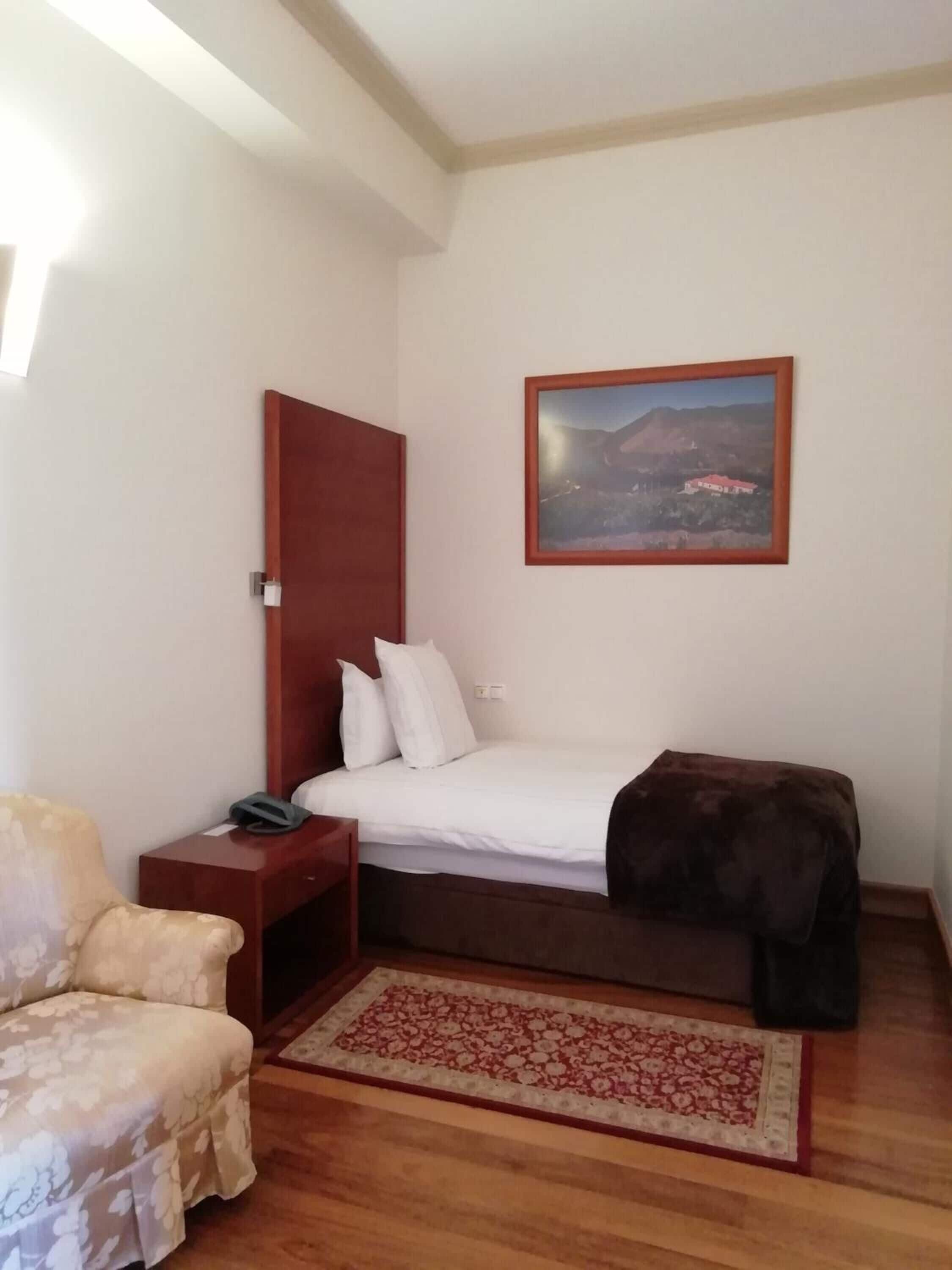 A bedroom with a bed, a nightstand, a chair, a wooden floor, a rug, and a framed picture on the wall.