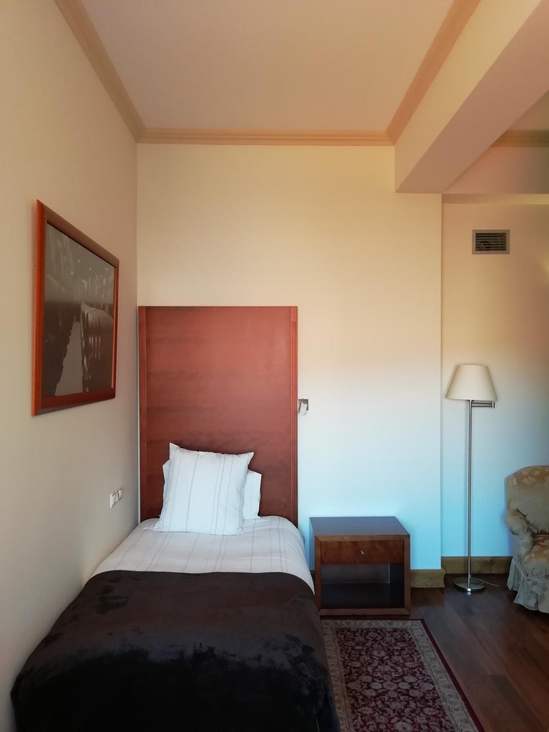 A bedroom with a wooden headboard, a bed with white linens, a nightstand, a floor lamp, and a chair.