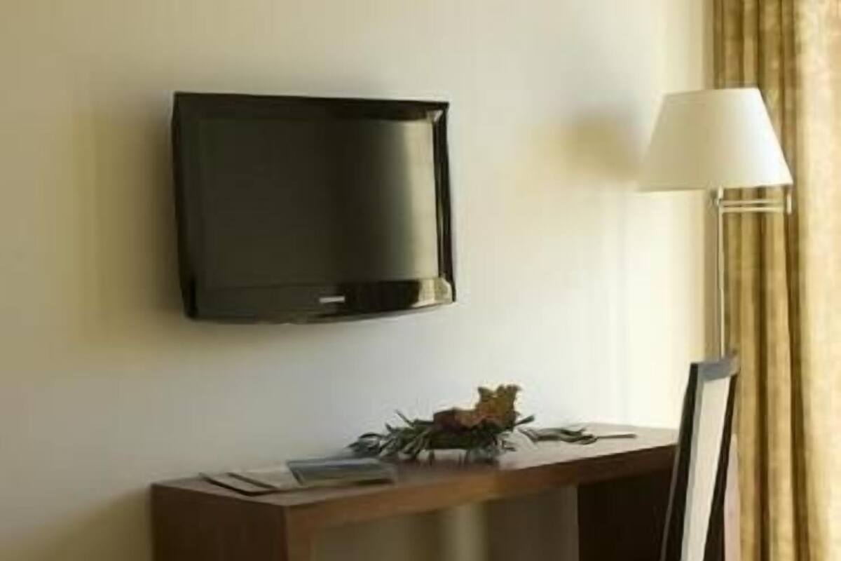 A hotel room with a flat-screen TV mounted on the wall, a desk with a chair, and a floor lamp.