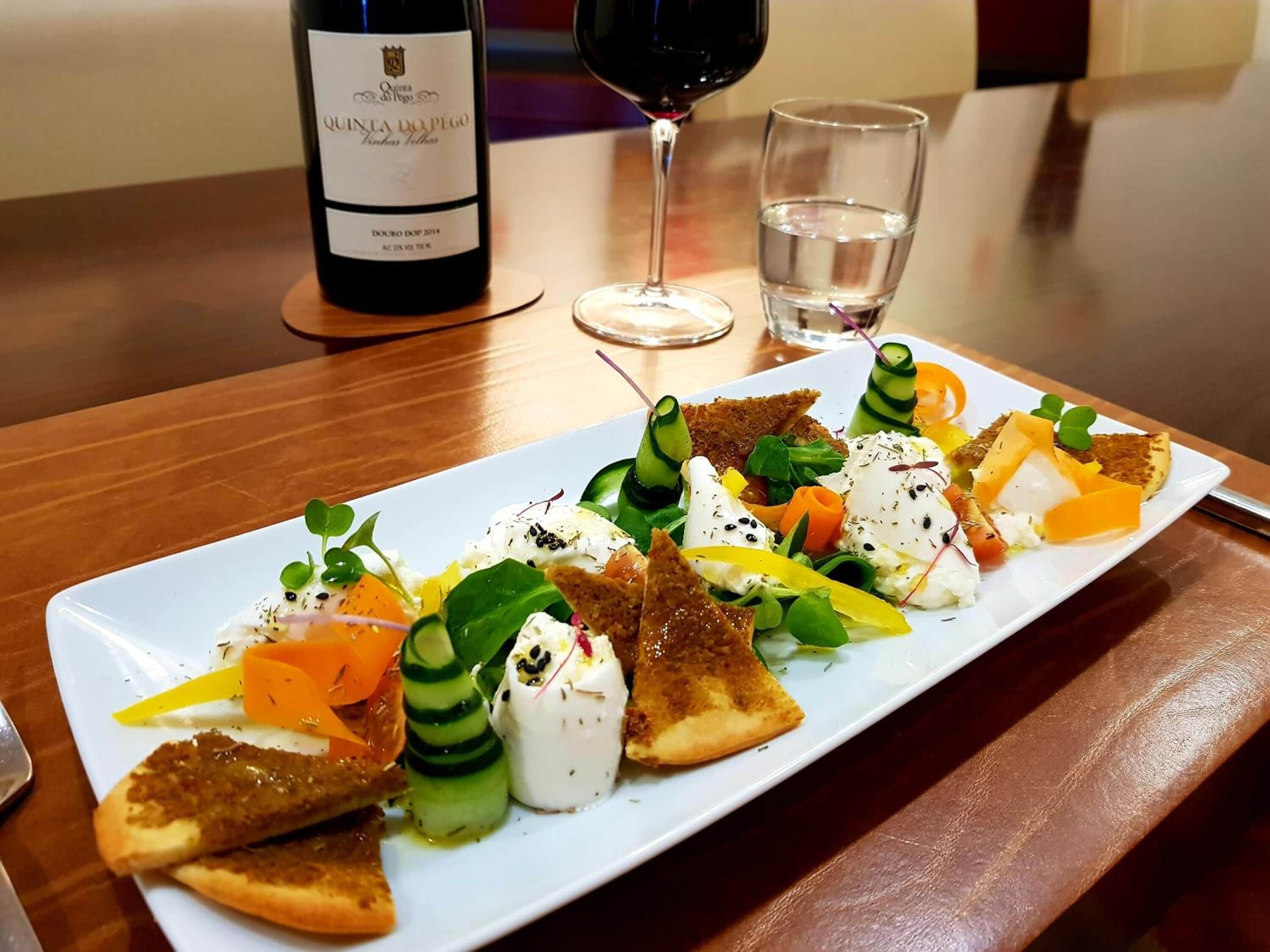 Un plato rectangular blanco con una variedad de aperitivos, incluyendo queso, vegetales y pan, acompañado de una botella de vino y una copa de vino tinto sobre una mesa de madera.
