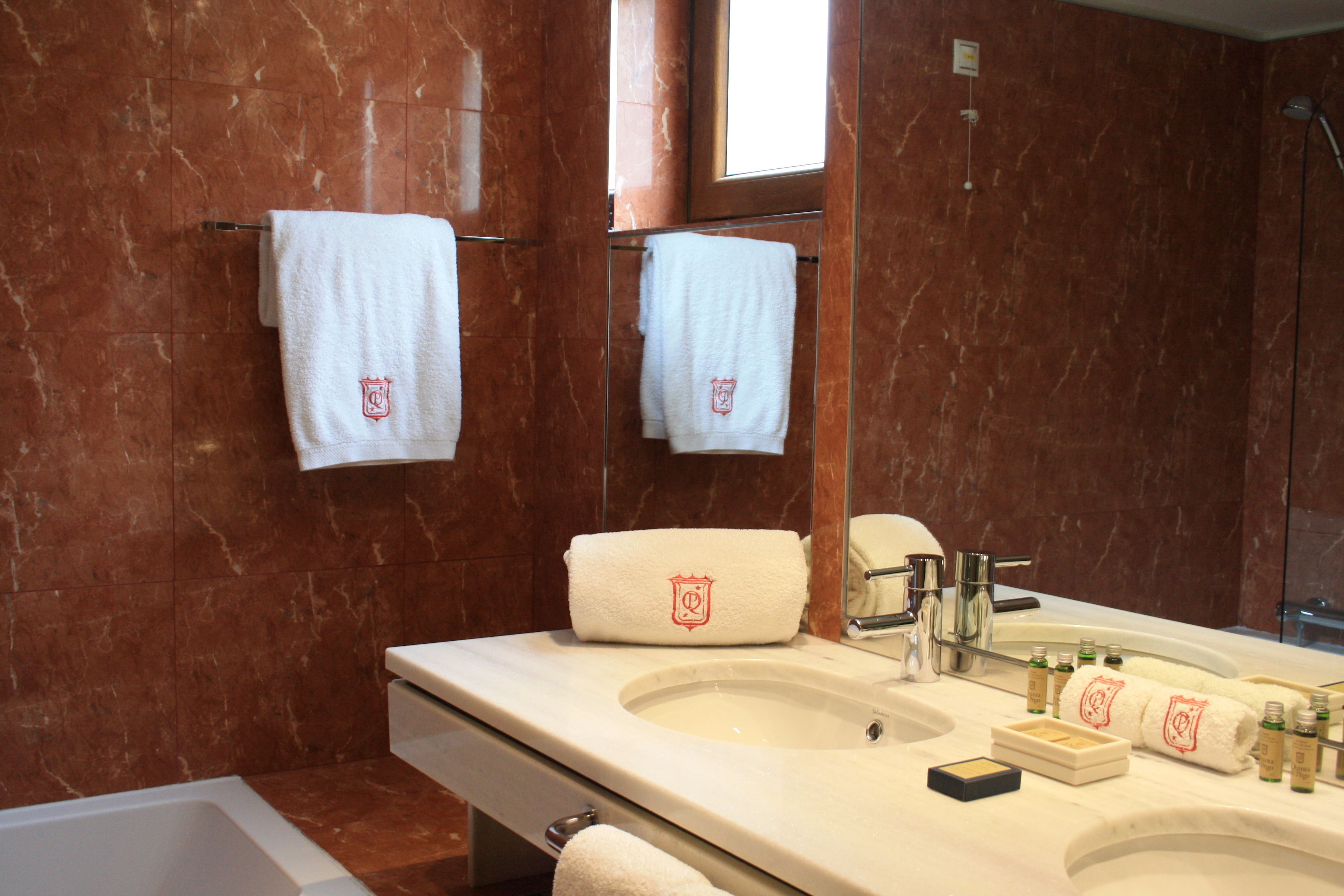 Un bagno con parete in marmo, un lavandino con articoli da toilette e due asciugamani appesi a ganci.