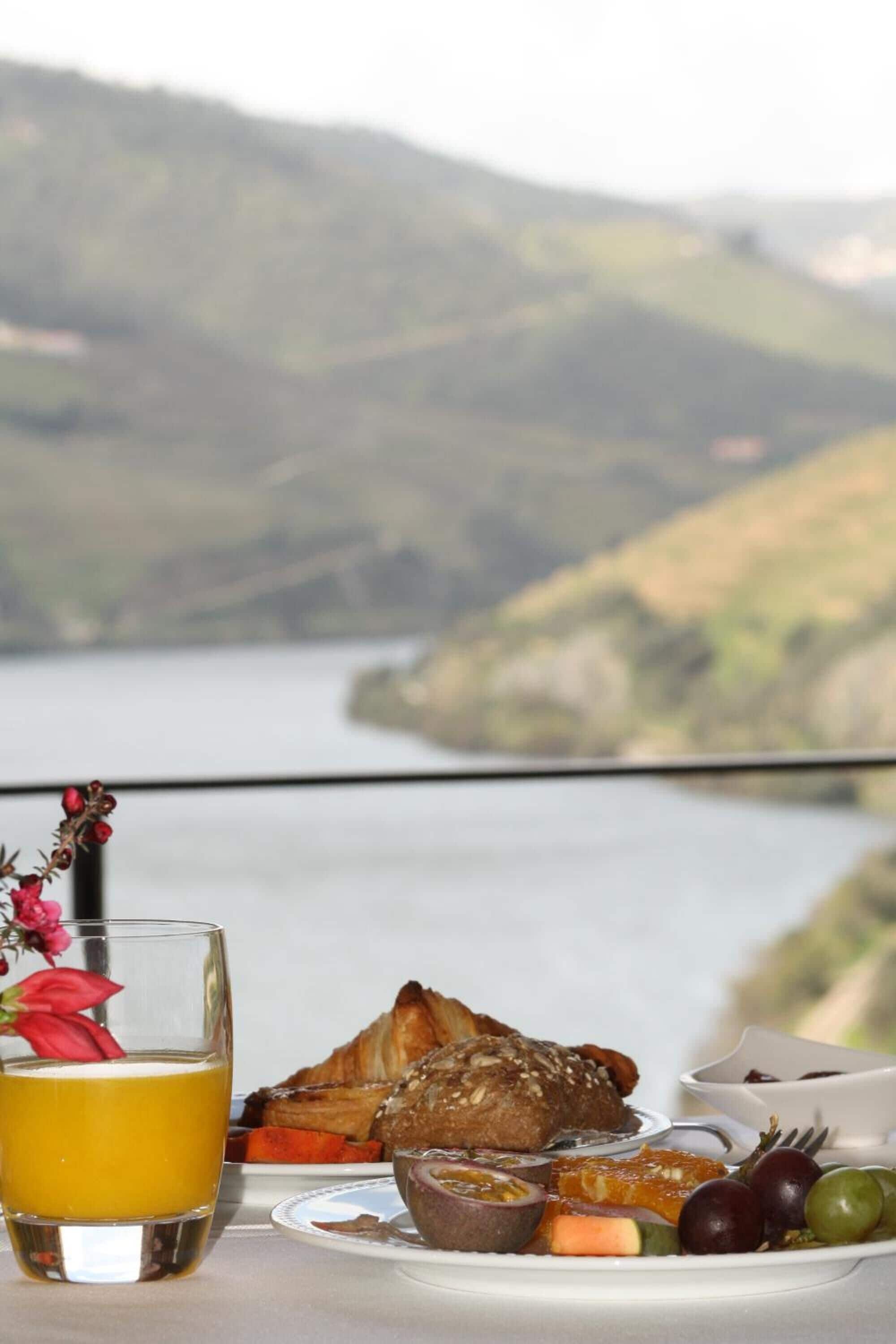 Desayuno con croissant, fruta y jugo, con vista a un lago y colinas.