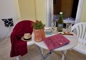 Una pequeña mesa redonda cubierta con un paño rojo, un sombrero, una planta en maceta, una botella de cerveza ZESI, un vaso, un cuaderno y un plato de comida.