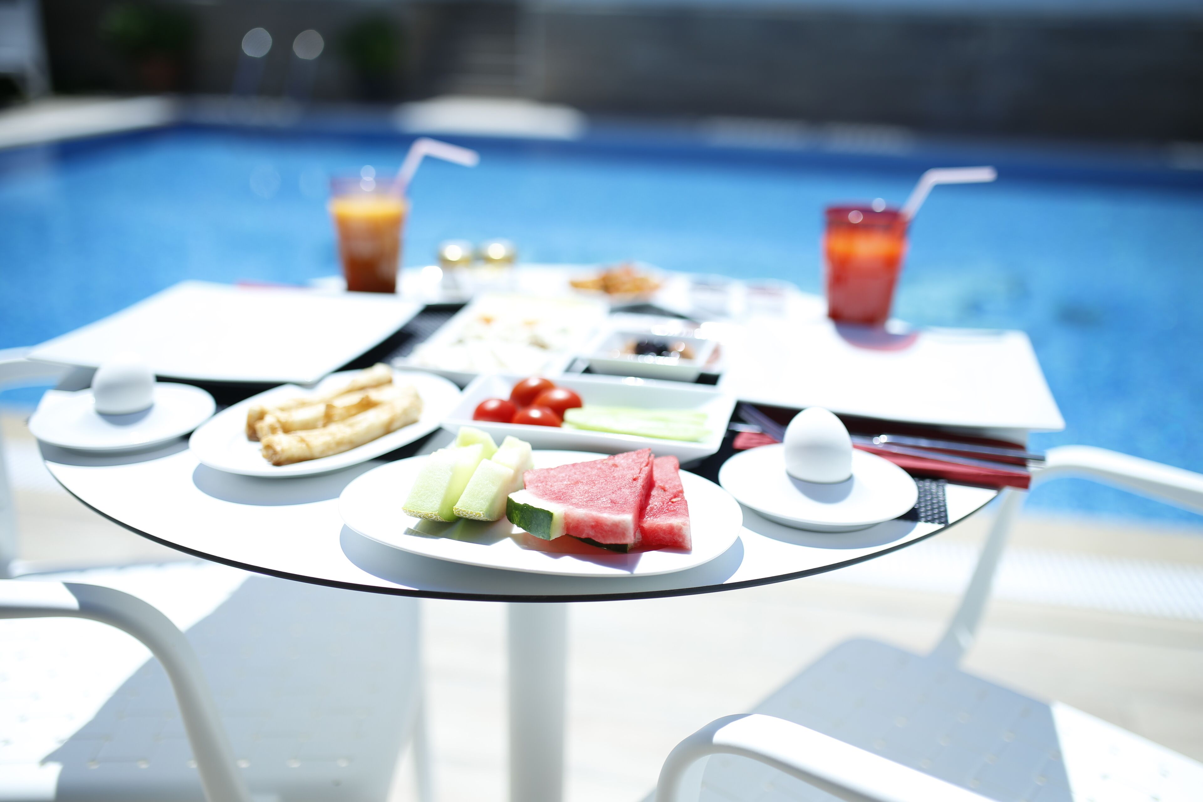 Una mesa con diversos alimentos, como sándwiches, frutas y bebidas, con una piscina al fondo.
