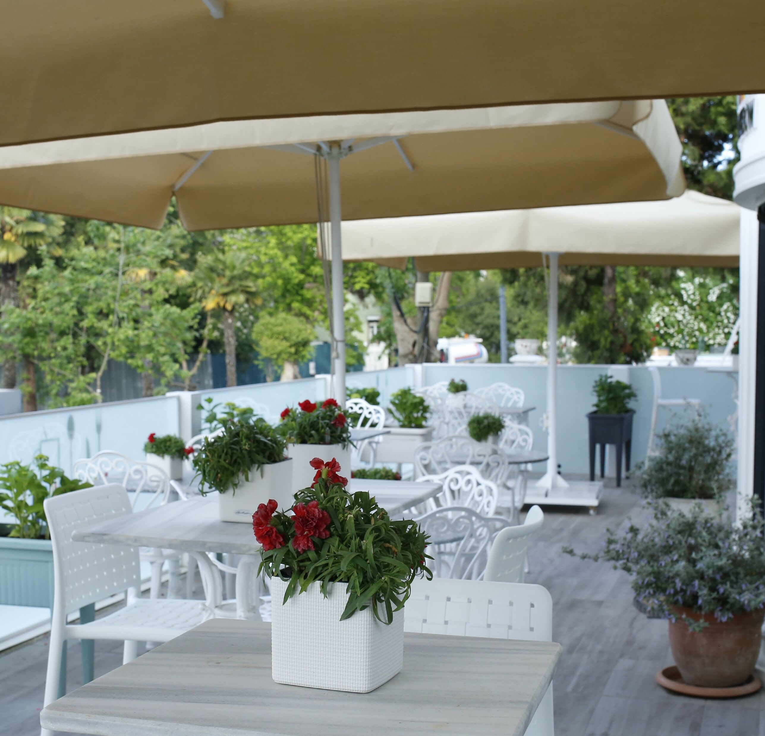 An outdoor patio area with white tables and chairs, covered by a large beige umbrella. There are potted plants and flowers on the tables.