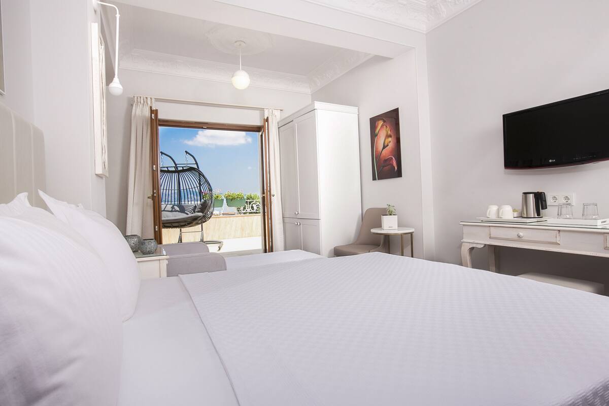 A hotel room with a large bed, a desk with a flat-screen TV, a chair, a small table, and a balcony with a view.