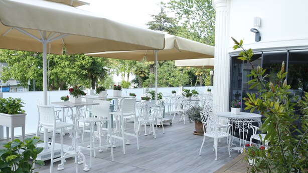 An outdoor patio with white metal chairs and tables, covered by beige umbrellas, surrounded by potted plants and a white fence. - Ascot Hotel Buyukada (Adalar)