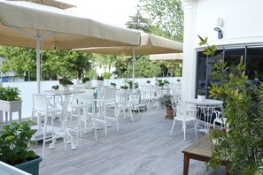 Un patio al aire libre con sillas y mesas de metal blanco, cubierto por sombrillas color beige, rodeado de plantas en macetas y una cerca blanca.