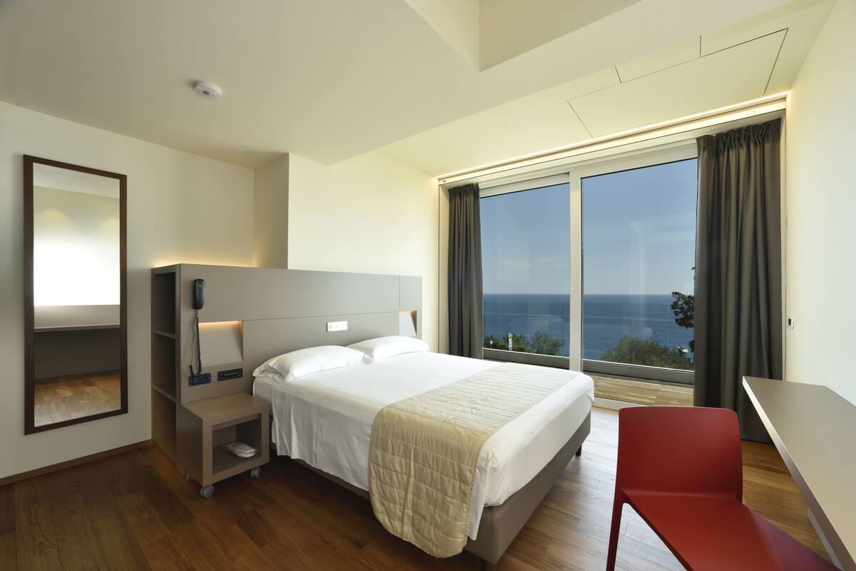A modern hotel room with a large bed, a desk, and a view of the sea.