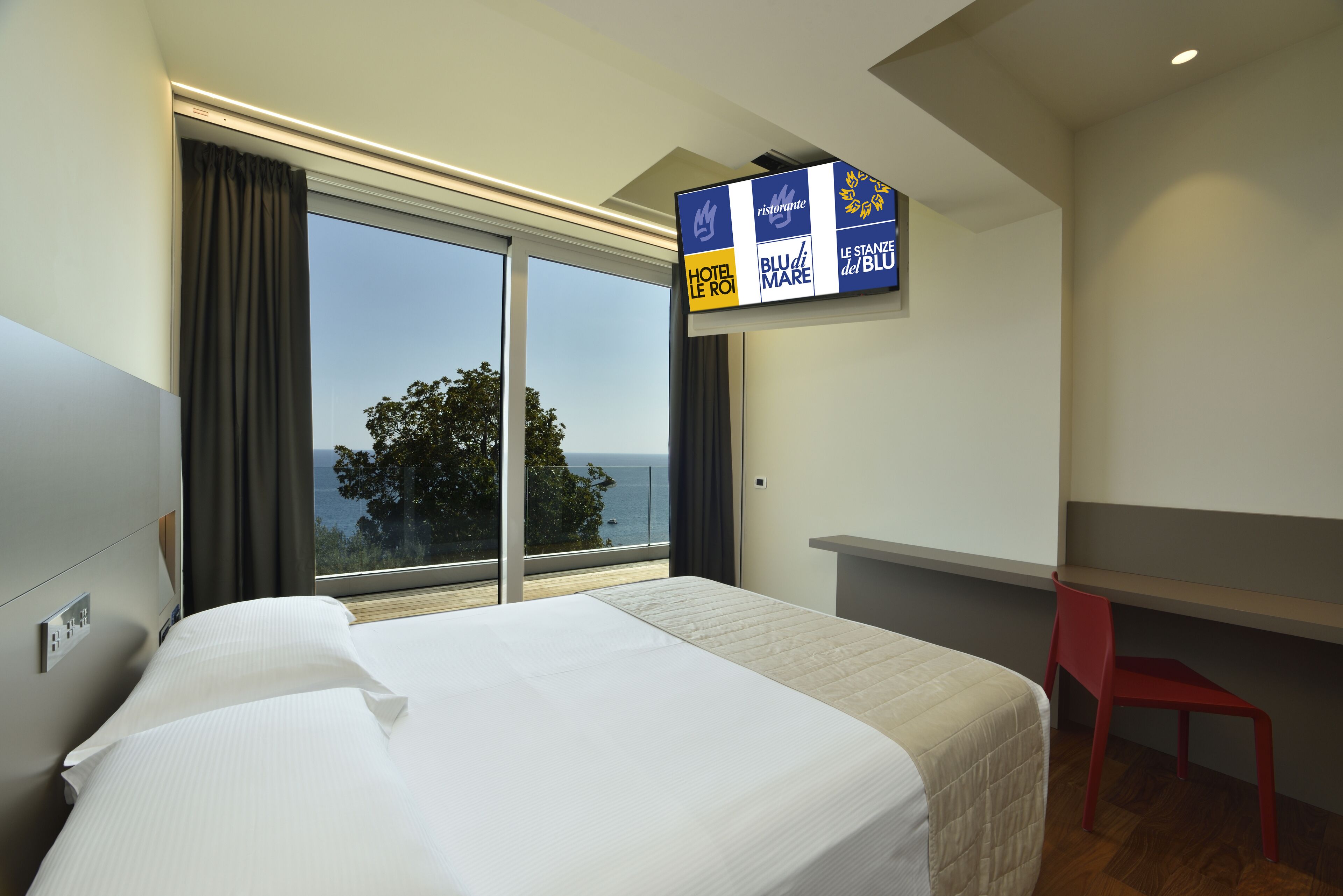 A hotel room with a large bed, a desk, and a TV displaying hotel names.