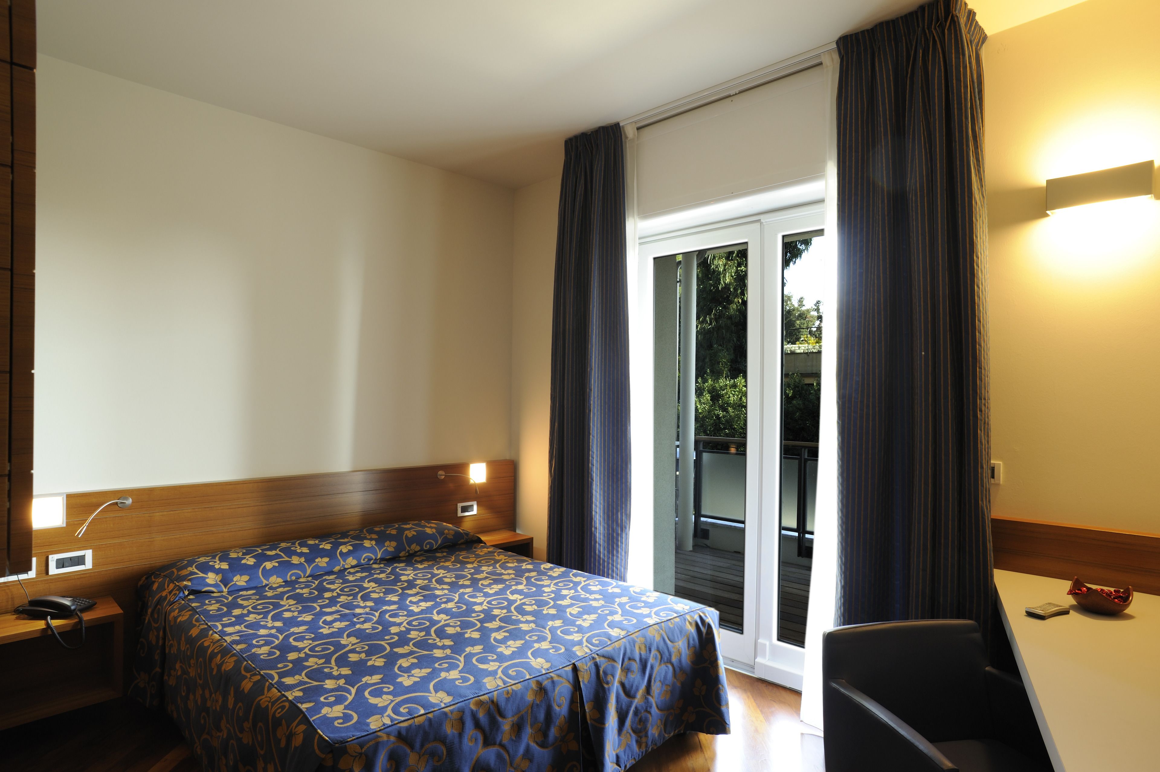 A hotel room with a large bed, a desk, a chair, a phone, and a window with curtains.