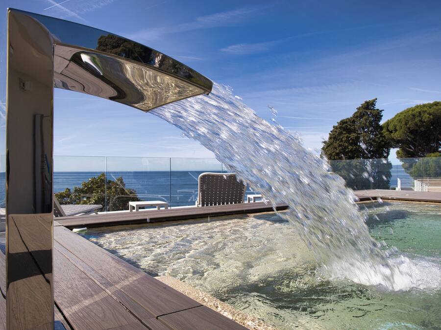 Una piscina con fuente, cubierta de madera, sillas de descanso y vista al mar.
