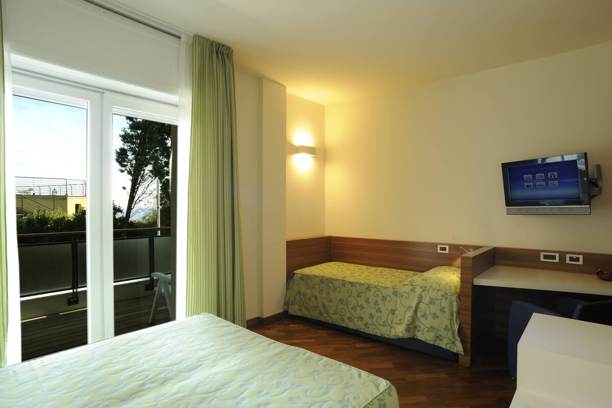 A hotel room with a bed, a desk, a chair, a TV, and a balcony with a view of the outside.