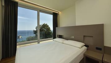 A modern hotel room with a large bed, a headboard with controls, a bedside table, and a view of the sea through large windows.