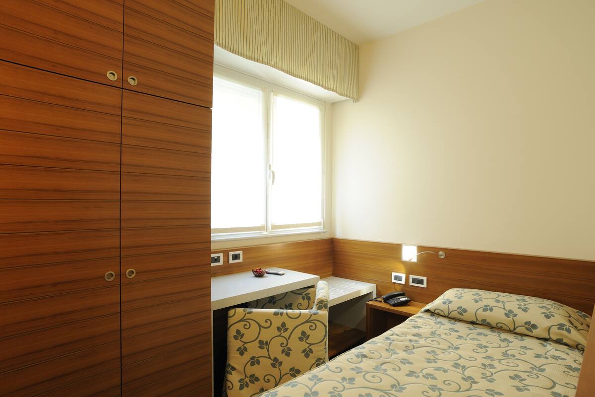 A compact room with a wooden wardrobe, a small desk with a chair, a bed with patterned bedding, and a window providing natural light.