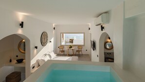 A modern hotel room with a large bathtub, a dining area with two chairs, a round mirror, and a view of the city.