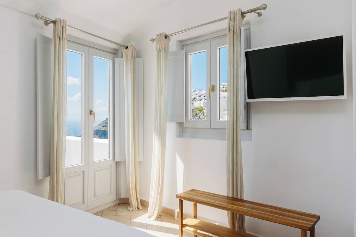 Una habitación con una cama, un banco de madera, un televisor montado en la pared y ventanas con cortinas.