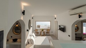 A modern hotel room with a bed, a round mirror, a small table with chairs, a TV, and a view of the sea.
