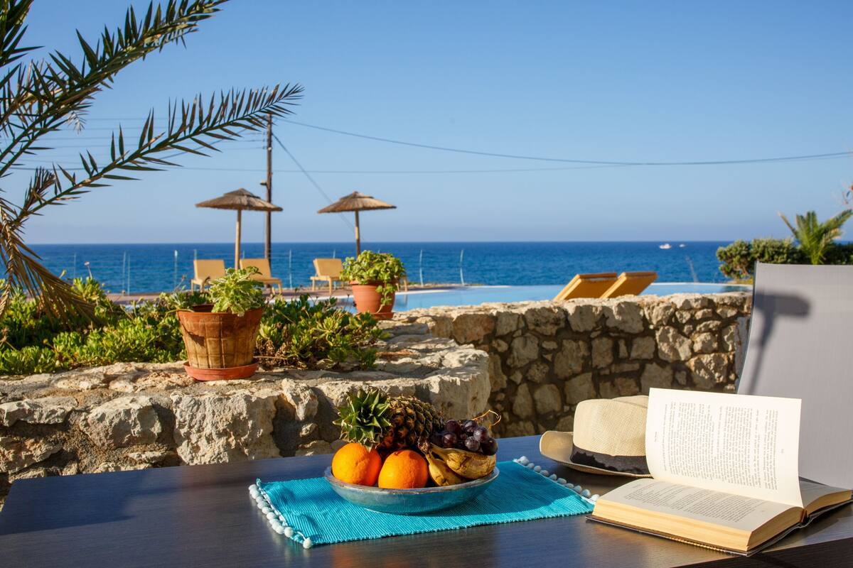 Ein Tisch mit Obstschale, einem offenen Buch und Blick auf das Meer und Strandstühle.