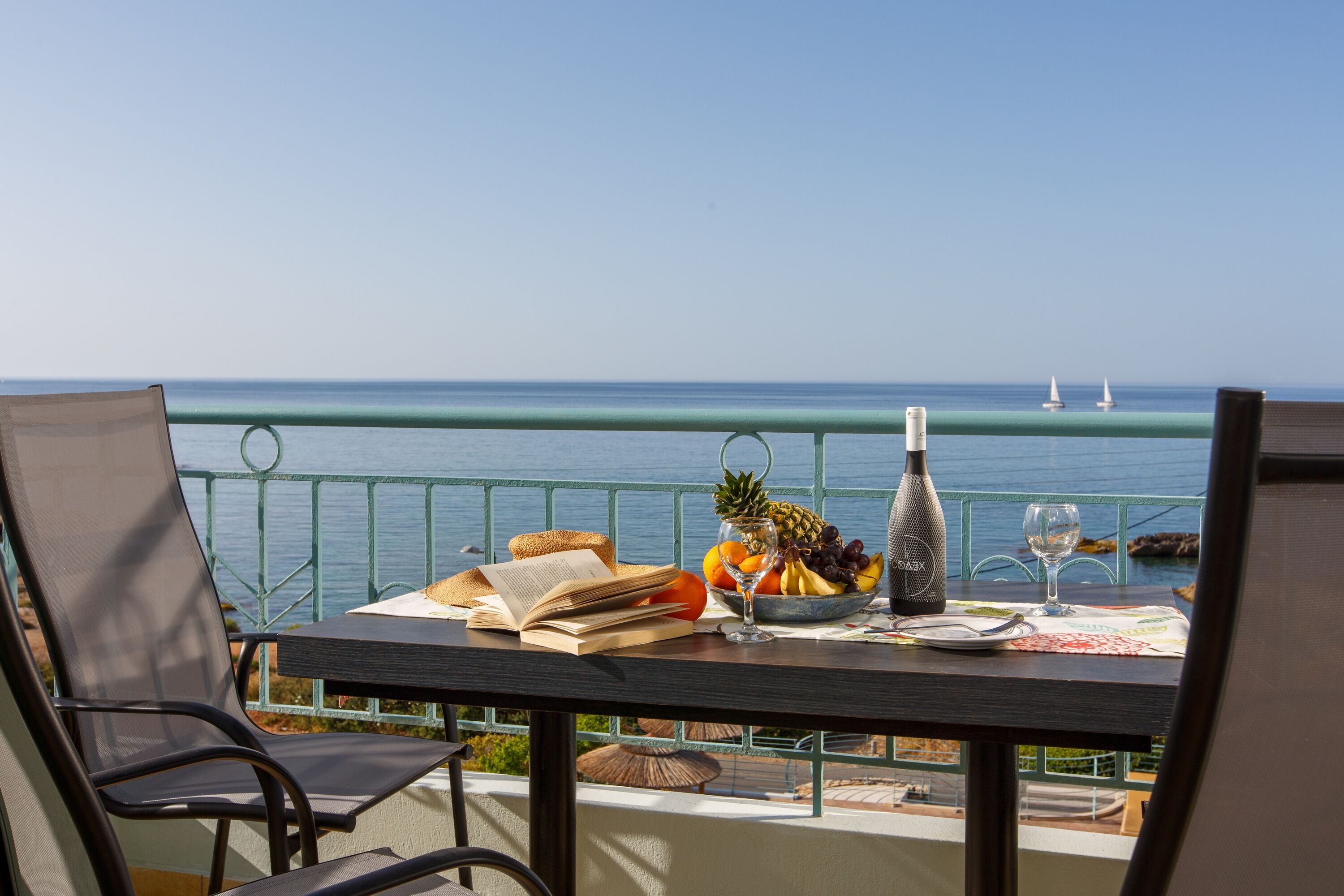 A balcony with a table set for a meal, overlooking the sea with sailboats in the distance.