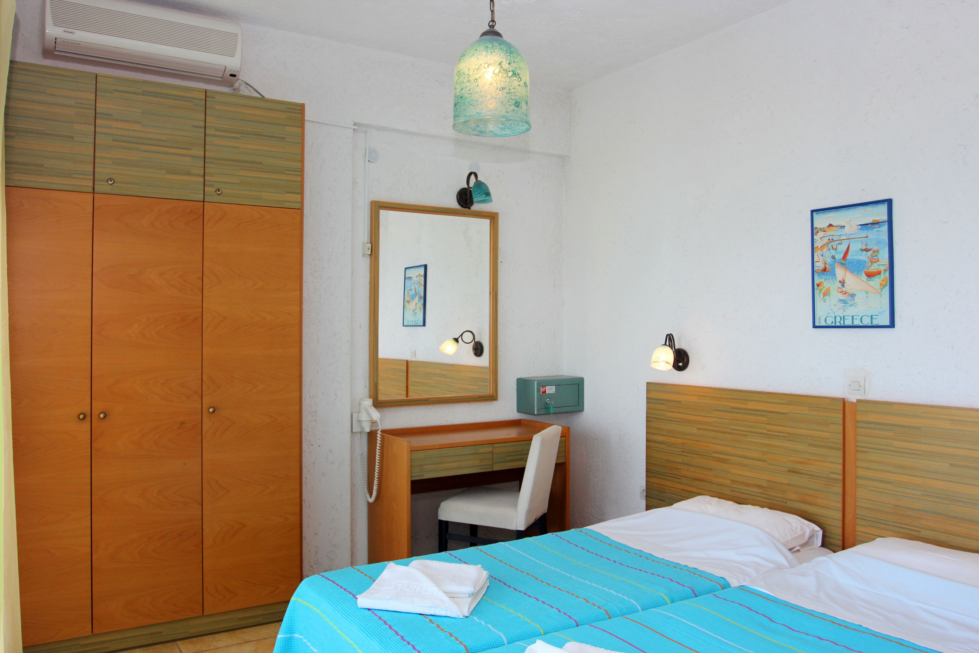 Une chambre d’hôtel comprenant un lit, un bureau, une chaise, une armoire, un miroir et un luminaire fixé au mur.