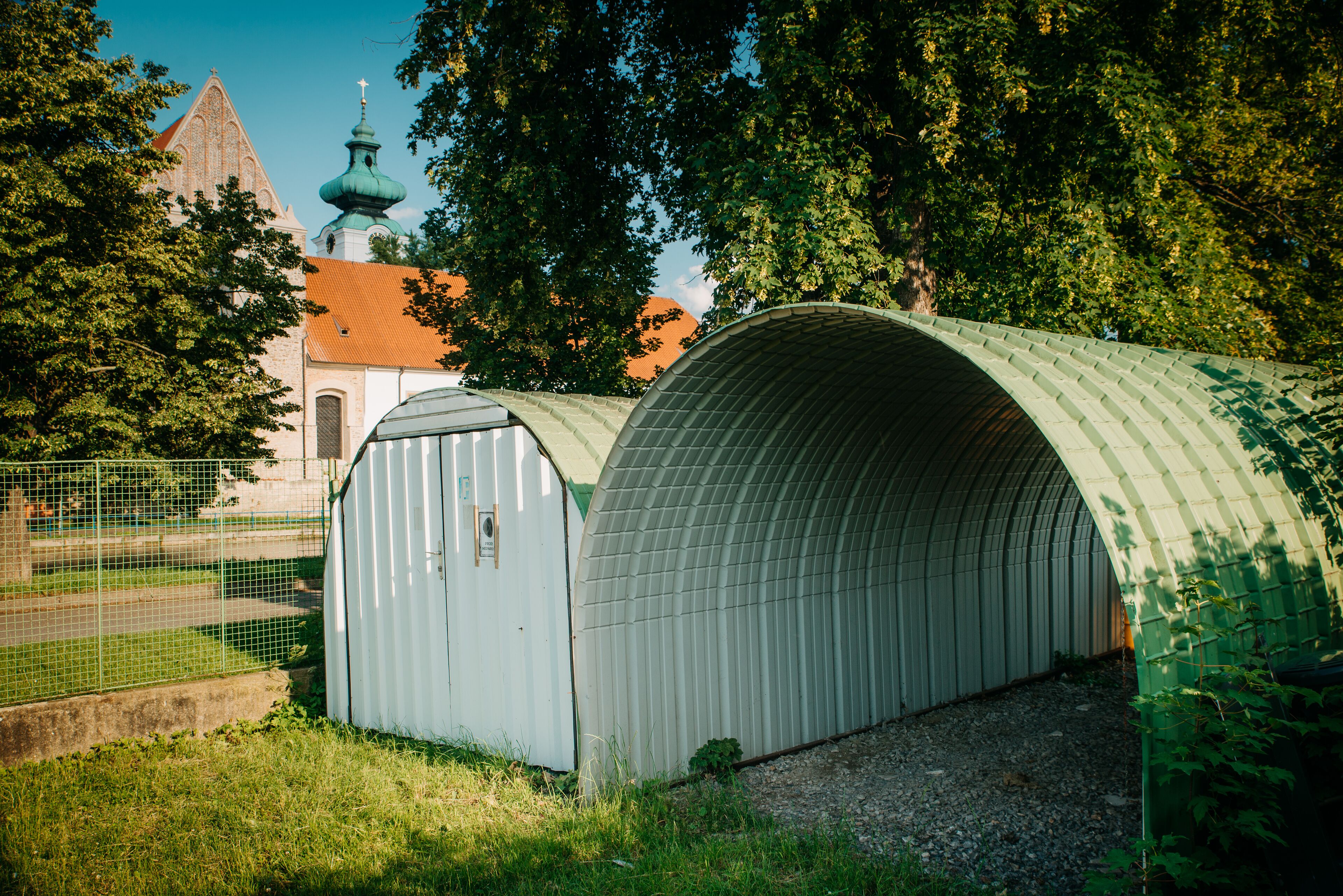 Een metalen constructie met een gebogen dak, mogelijk een opslag- of bijgebouw, naast een kerk met een groene koepel en een kruis.