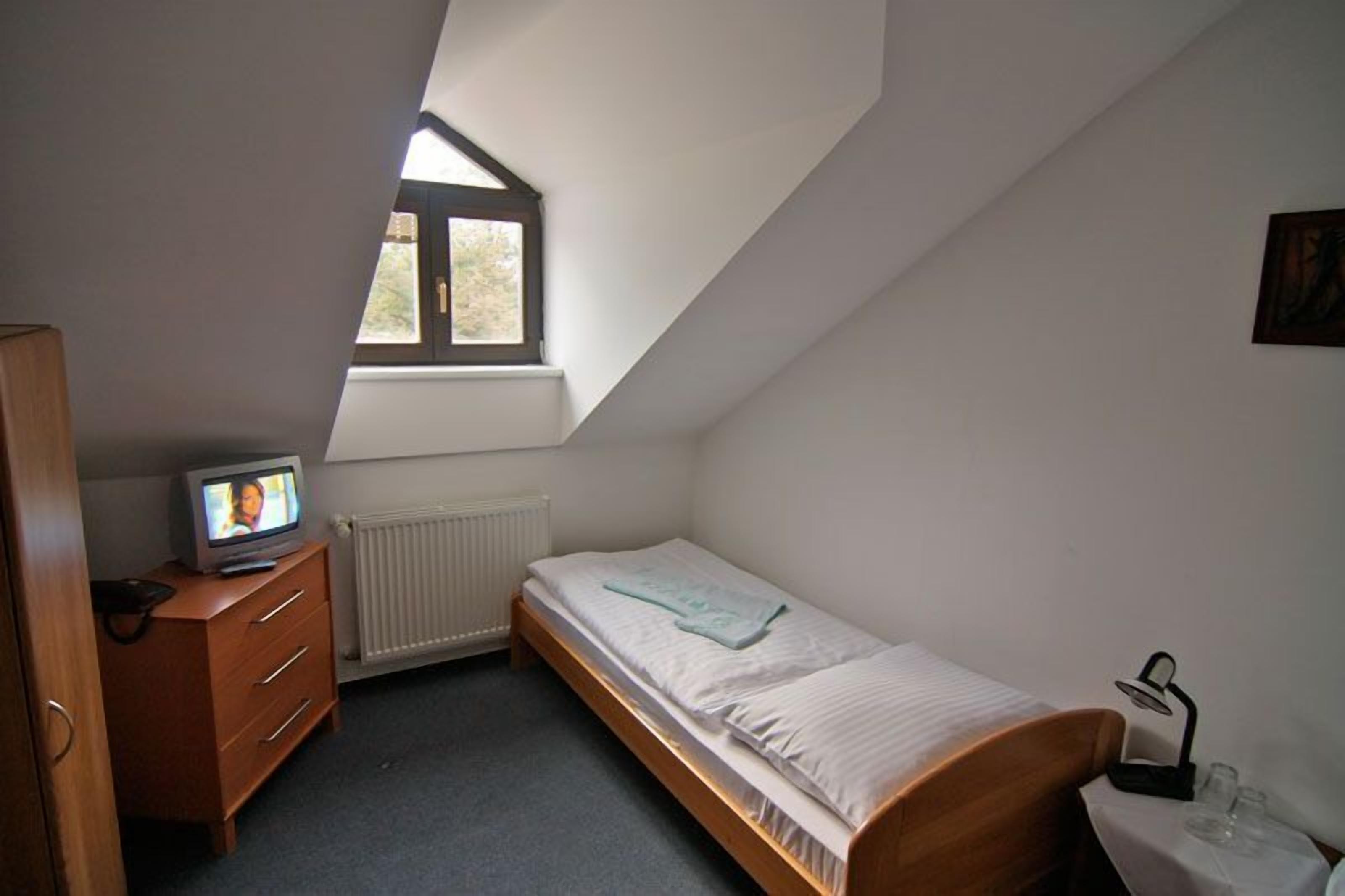 Una habitación pequeña con cama individual, techo inclinado, ventana, radiador, cómoda de madera, televisor y mesita de noche con lámpara.