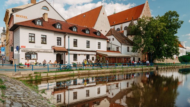 A hotel with multiple stories, a red-tiled roof, and a reflection in the water. - Hotel Klika (Ceske Budejovice)