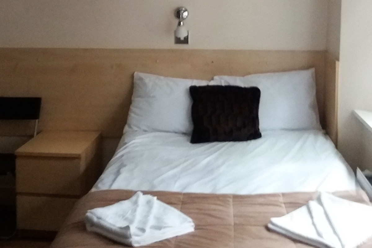 Ein Hotelzimmer mit Bett, Flachbildfernseher an der Wand, einem Bild über dem Bett und einem Nachttisch mit Lampe.