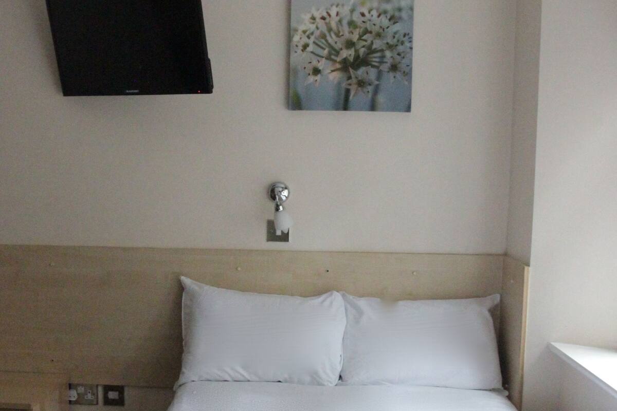 A hotel room with a bed, a flat-screen TV mounted on the wall, a painting above the bed, and a bedside table with a lamp.