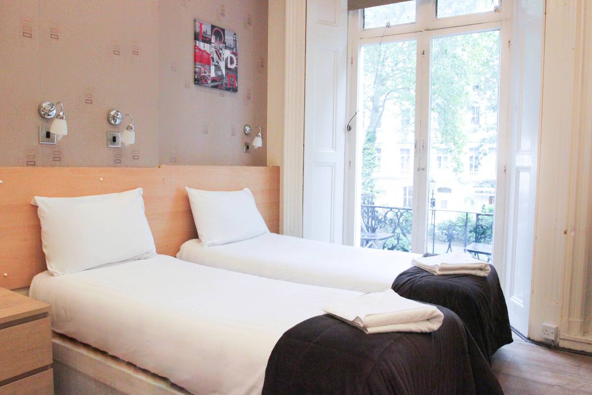 A hotel room with two beds, a wooden headboard, a wall-mounted light fixture, and a large window with a view of the outside.