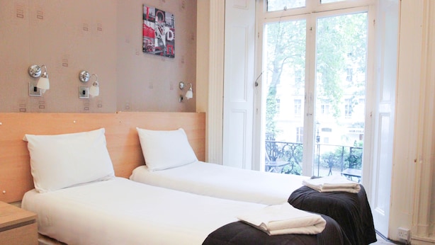 A hotel room with two beds, a wooden headboard, a wall-mounted light fixture, and a large window with a view of the outside. - Tony's House Hotel (London)