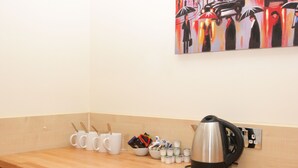 A kitchen counter with coffee mugs, a kettle, and a painting of people with umbrellas. - Tony's House Hotel (London)