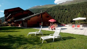 A wooden lodge with a patio area, outdoor seating, and a mountainous backdrop. - Hotel Santa Caterina (Valfurva)
