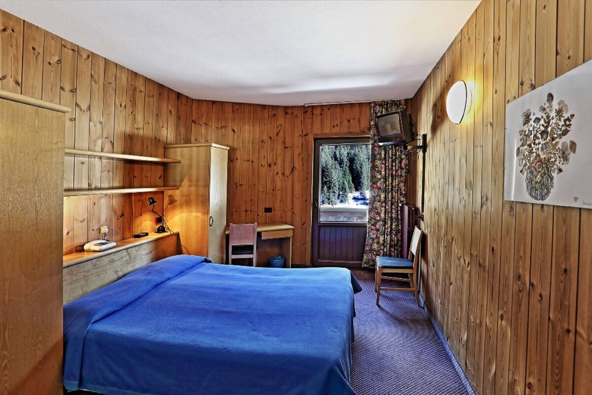 A wooden-paneled room with a bed, desk, chair, and a window with a view of greenery.