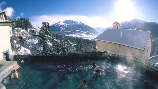 A hot spring with clear water, surrounded by snow-covered mountains and a building with a sloped roof. - Hotel Santa Caterina (Valfurva)