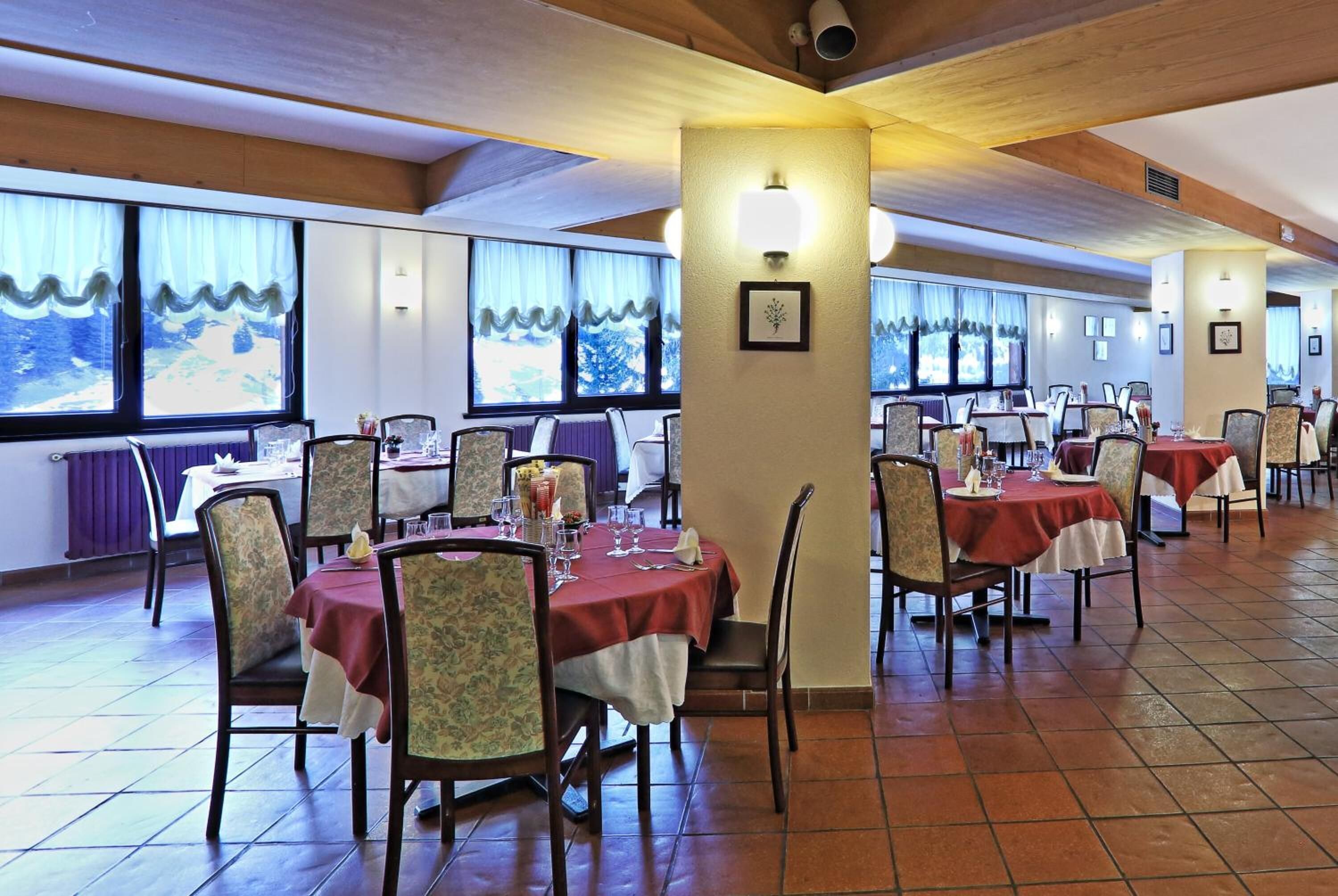 A dining area with multiple tables and chairs, large windows with curtains, and wall-mounted light fixtures.