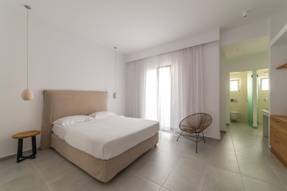 A modern bedroom with a large bed, a small side table, a chair, and a view of the bathroom through an open door.