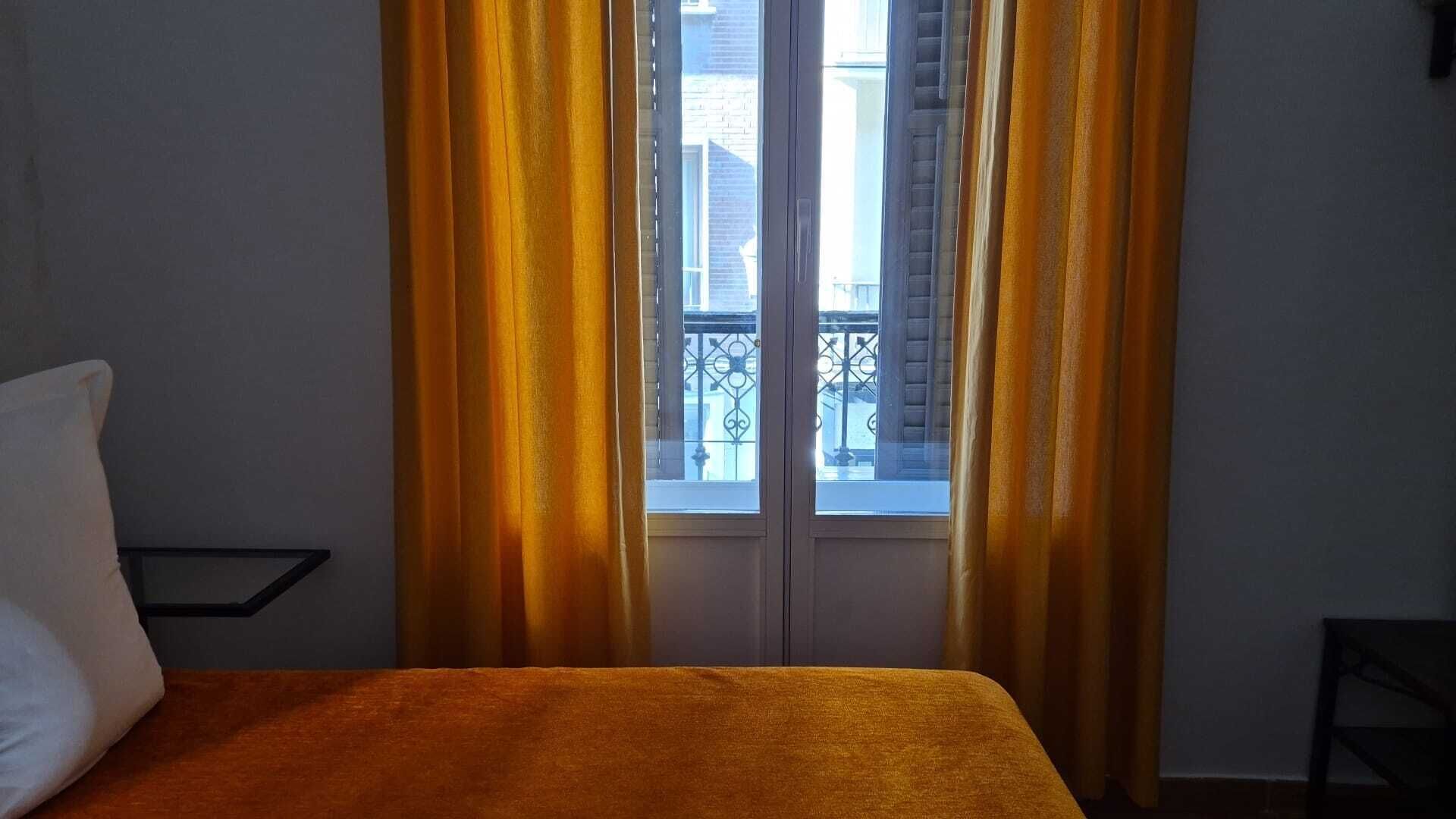 A room with a bed, a window with yellow curtains, and a view of buildings outside.
