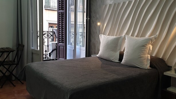 Un dormitorio con cama, almohadas, una mesita de noche, ventana y balcón.
