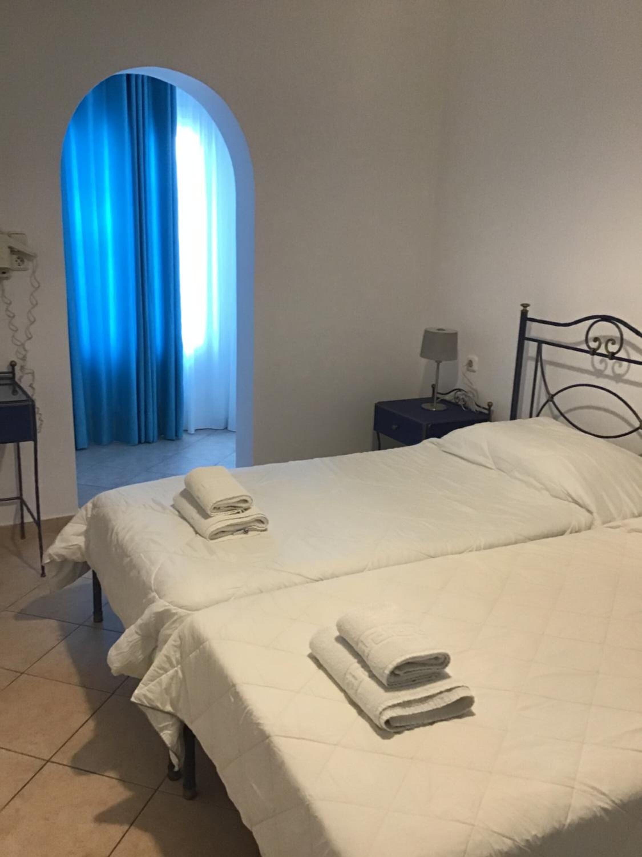 Habitación de hotel con dos camas, una mesita de noche con lámpara, cortina azul y suelo de baldosas.