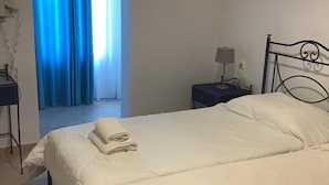 A hotel room with two beds, a nightstand with a lamp, a blue curtain, and a tiled floor. - Hotel Lignos (Santorini)